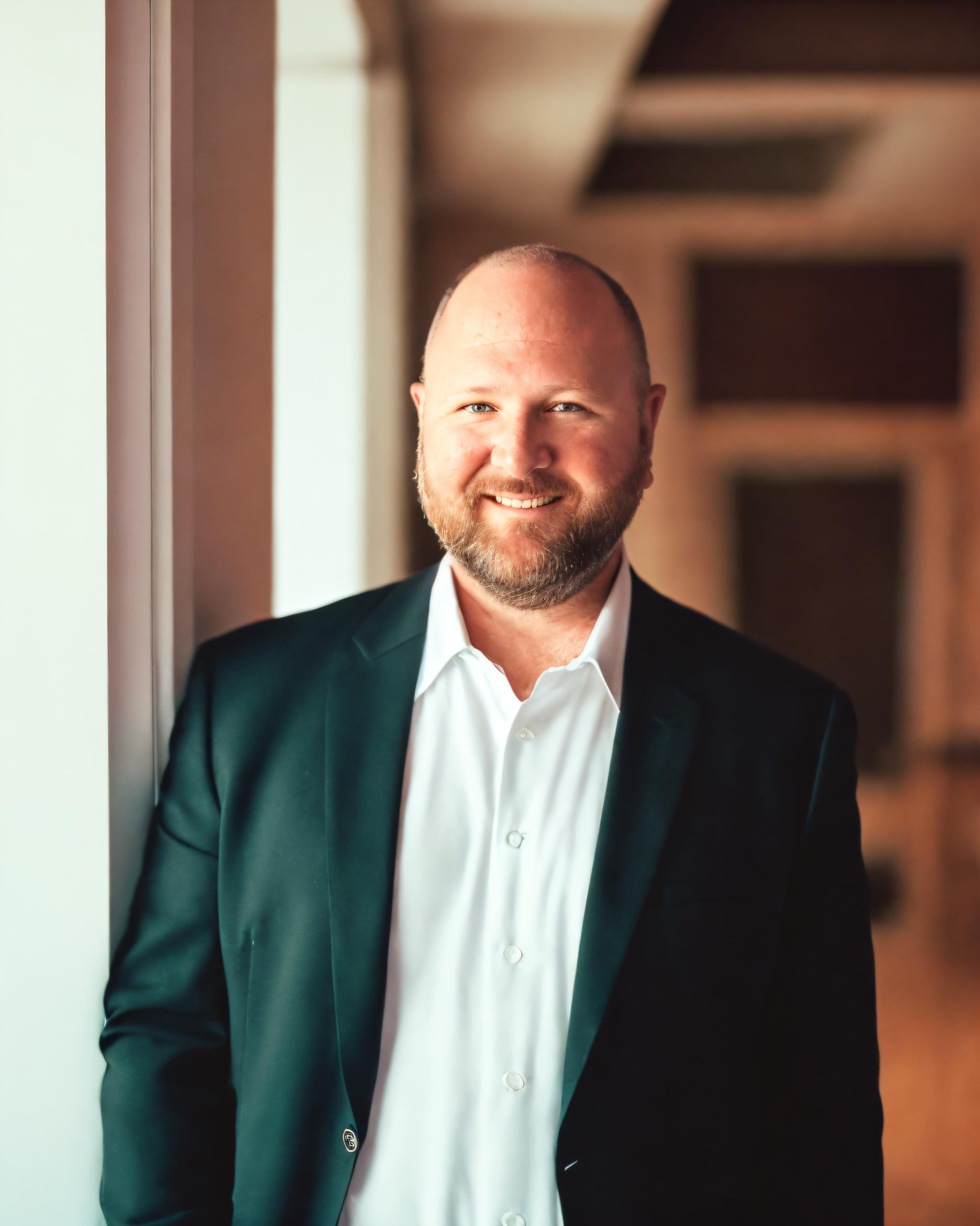 A man with a beard and shaved head, wearing a dark suit jacket and white dress shirt, stands smiling next to a window in a softly lit, modern indoor setting.